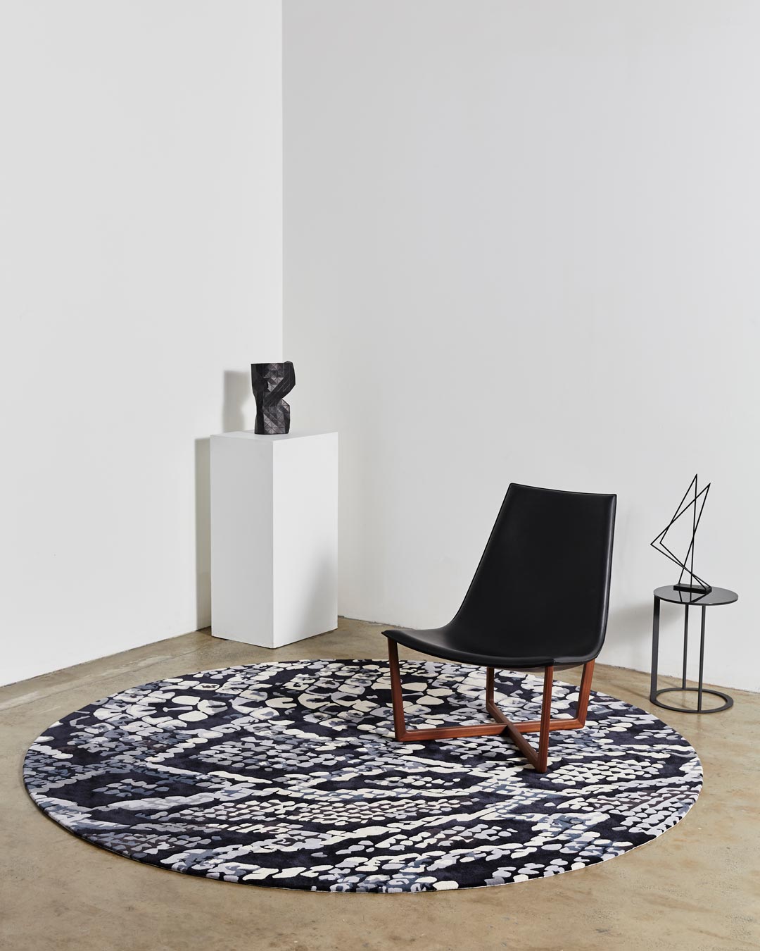 A round, patterned rug with a floral design in shades of blue and black, placed on a floor with a chair, a lamp, and a small wooden stand with a black sculpture on top.